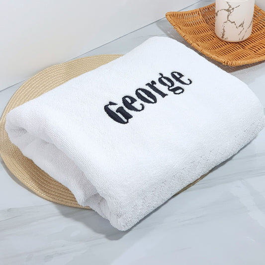 White bath towel with 'George' embroidered on it, placed on a woven mat.