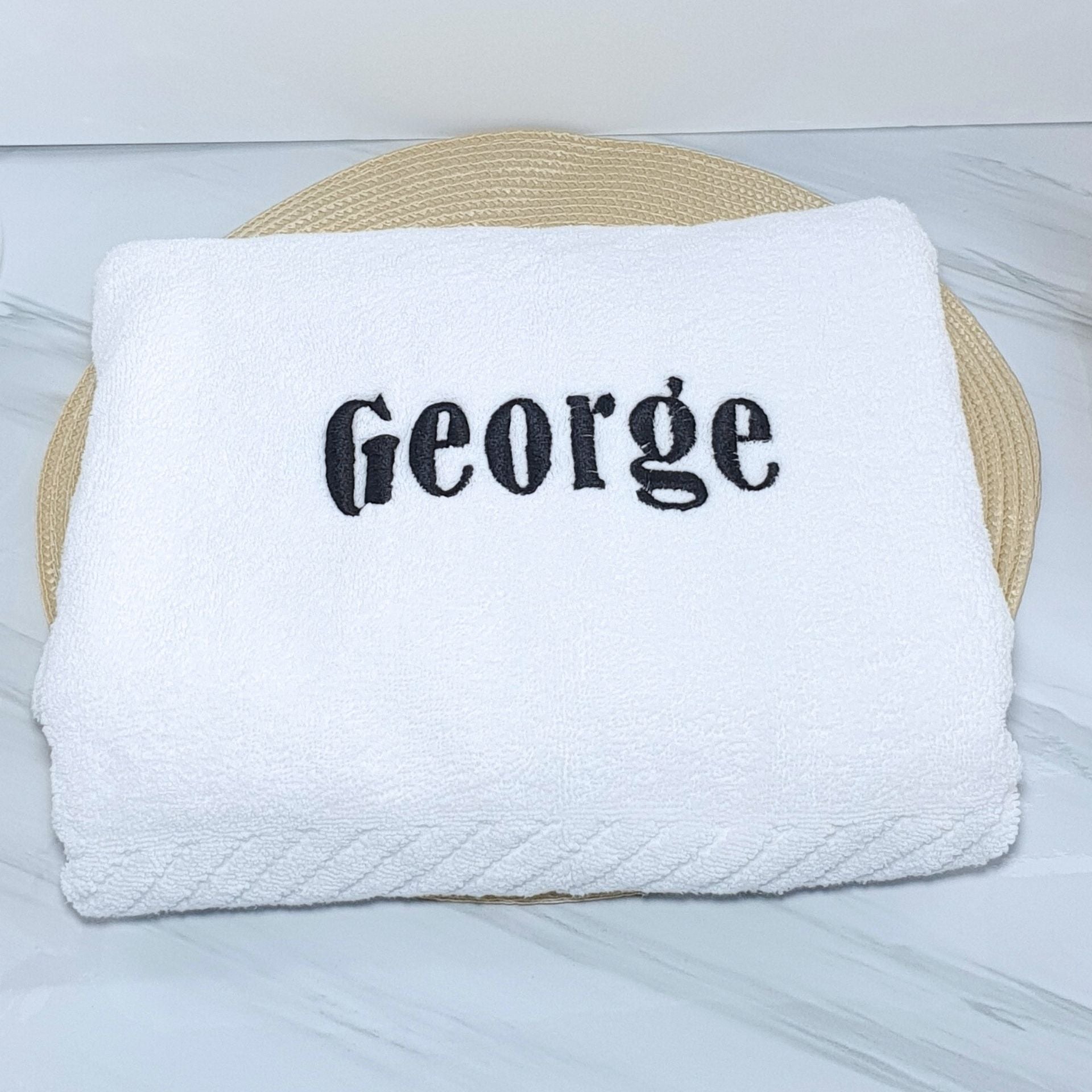 White bath towel with 'George' embroidered on it, placed on a woven mat.