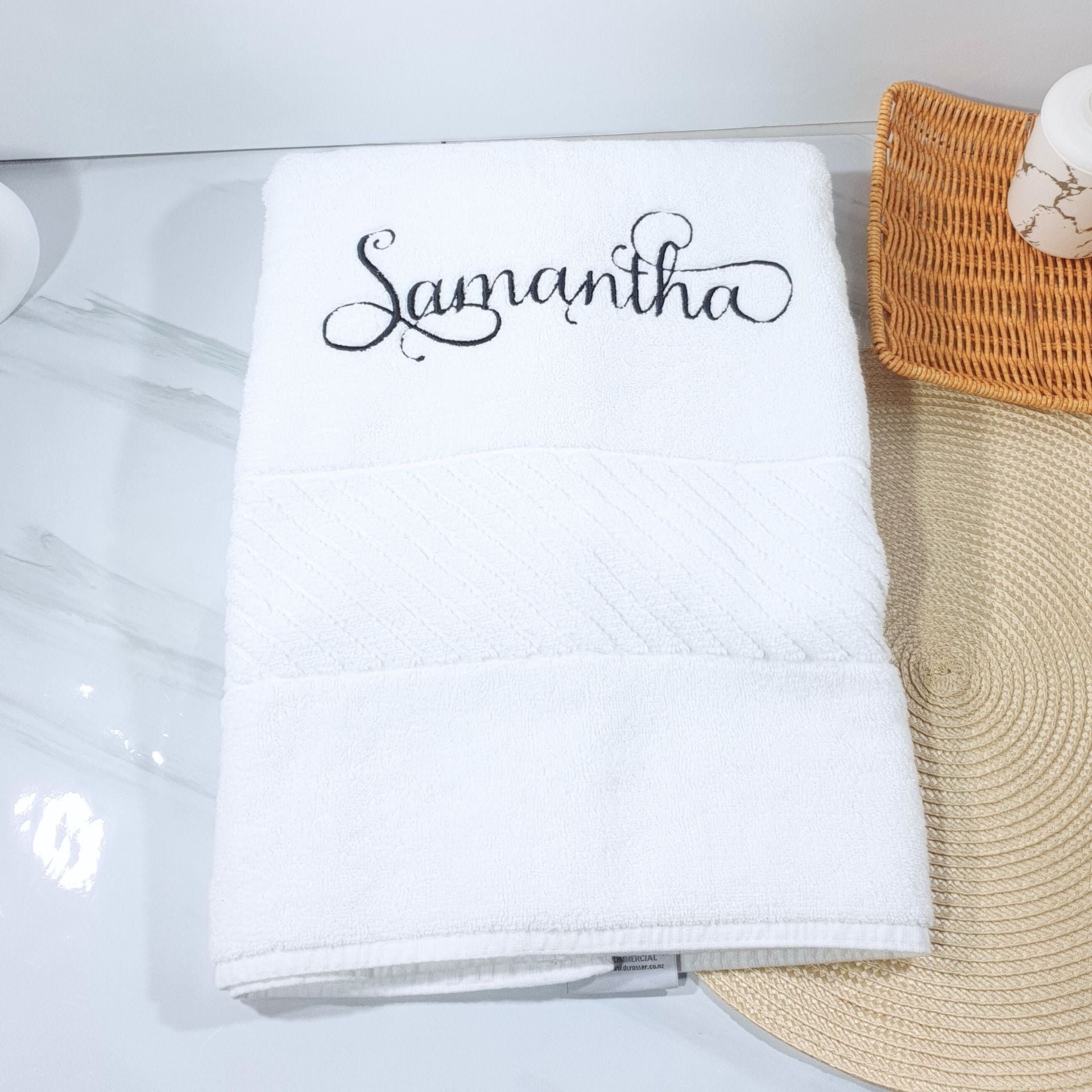 White bath towel with 'Samantha' embroidered on it, placed on a marble surface with a woven mat.