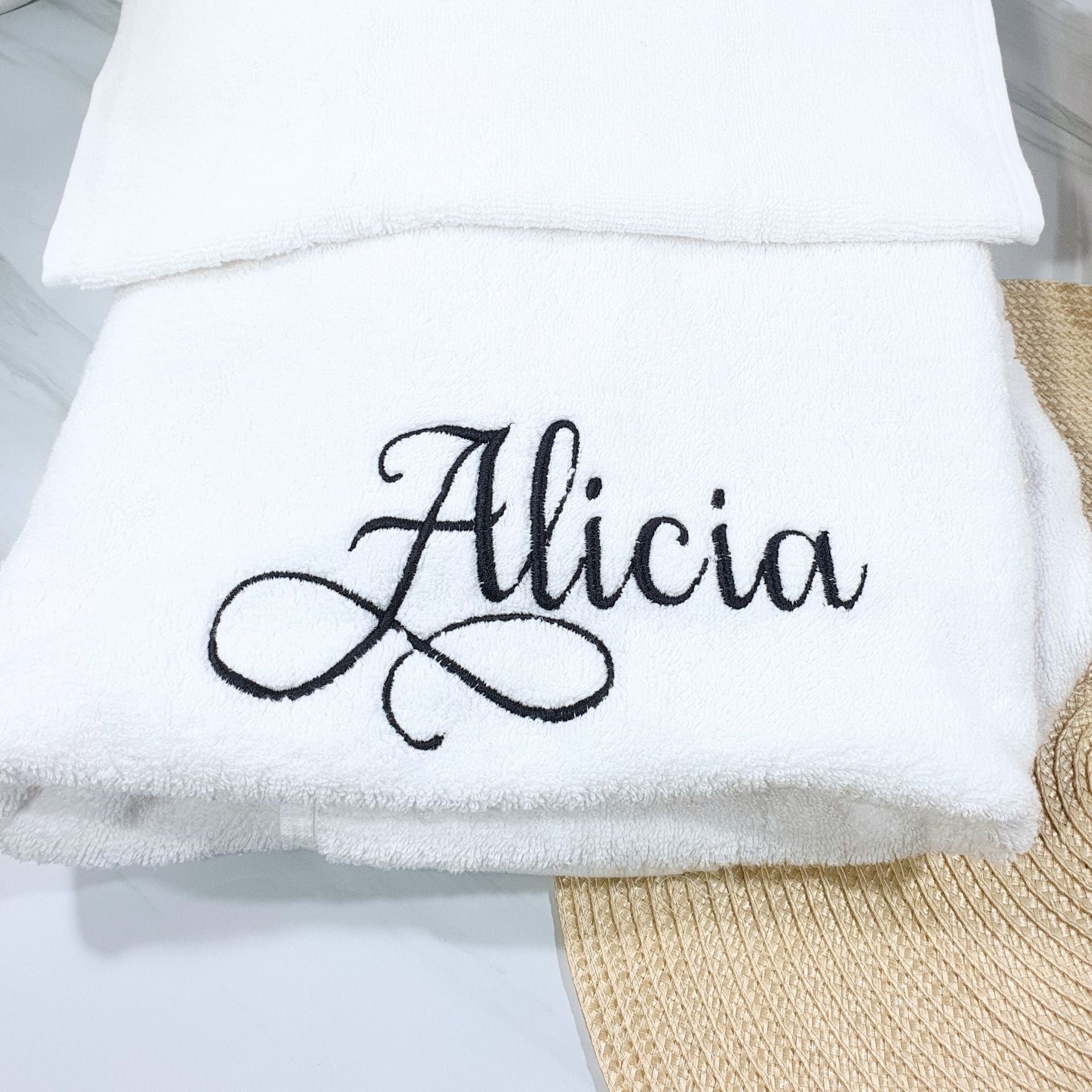 White bath towel with 'Alicia' embroidered on it, placed on a textured surface.