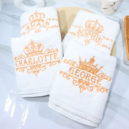 Set of white towels with embroidered names and crowns on a light gray surface.
