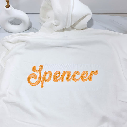 White hoodie with 'Spencer' embroidered on it, lying on a white surface.